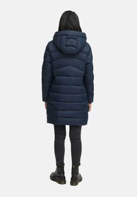 Mujer vestida con un abrigo acolchado largo de color azul marino con capucha, jeans negros ajustados y botas negras con cordones, de pie de espaldas.