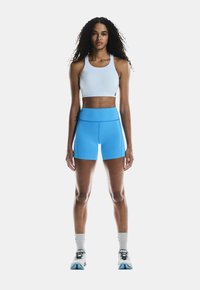 Top sportivo azzurro chiaro con scollatura rotonda abbinato a pantaloni corti alti di colore blu vivace. Il modello indossa scarpe da atletica bianche e blu con calzini grigi.
