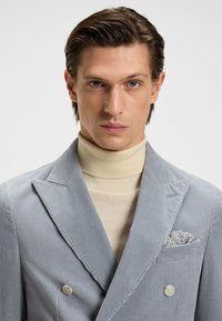 Blazer doppiopetto in corda azzurro chiaro con revers a spacco, abbinato a un dolcevita beige e a un fazzoletto da taschino a fantasia.