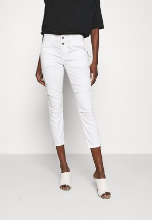 Kobieta ubrana w krótkie białe spodnie z bocznymi kieszeniami na zamek, czarny top oraz białe odkryte mulesy na obcasie, stojąca na jednolitym tle.