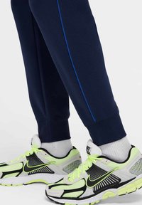 Granatowe spodnie dresowe z niebieskimi bocznymi paskami w połączeniu z czarnymi i neonowo-żółtymi sneakersami z siateczką i gumowymi detalami.