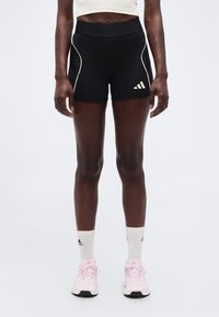 Αθλητικό άτομο που φοράει μαύρο σορτς Adidas, άσπρες κάλτσες Adidas και ροζ αθλητικά παπούτσια Adidas, στέκεται σε απλό φωτεινό φόντο.