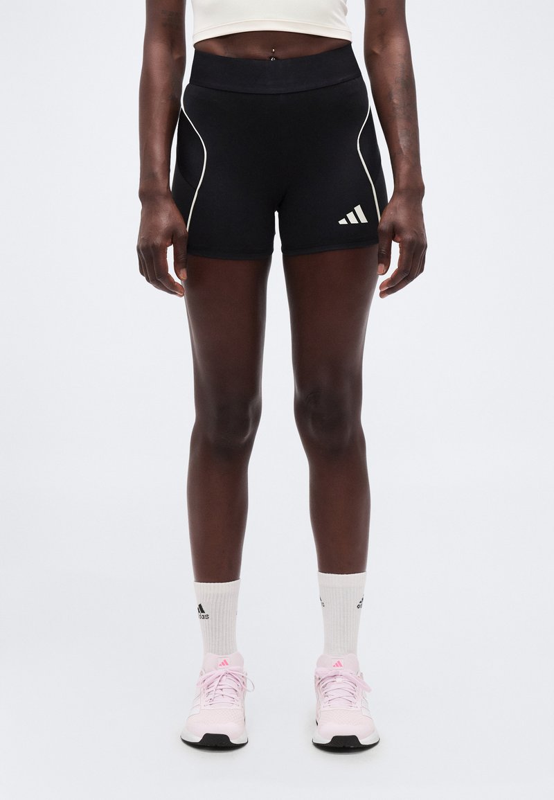 Αθλητικό άτομο που φοράει μαύρο σορτς Adidas, άσπρες κάλτσες Adidas και ροζ αθλητικά παπούτσια Adidas, στέκεται σε απλό φωτεινό φόντο.