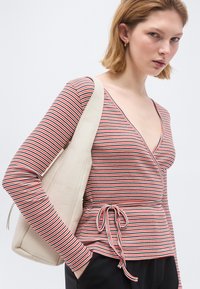 Γυναίκα που φοράει κόκκινο, λευκό και μαύρο ριγέ τοπ τύπου wrap με μακριά μανίκια, μαύρο παντελόνι, χρυσούς κρίκους στα αυτιά και κρατά μια μπεζ τσάντα ώμου.