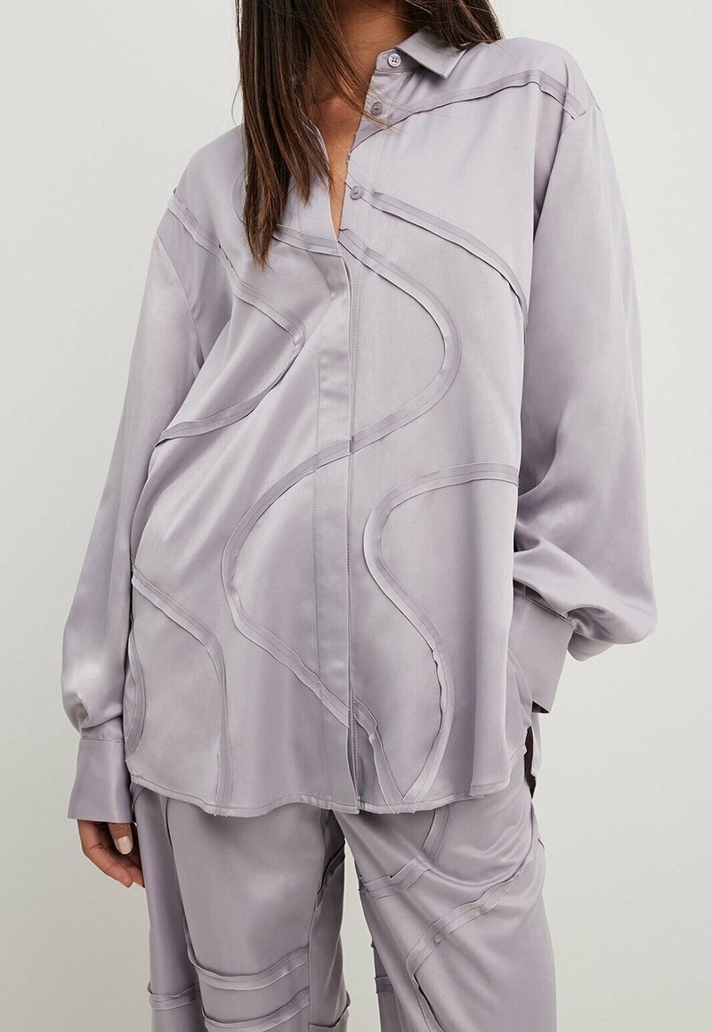 Femme portant une chemise ample violet clair et un pantalon avec des détails de coutures courbes sur le tissu, sur un fond uni.