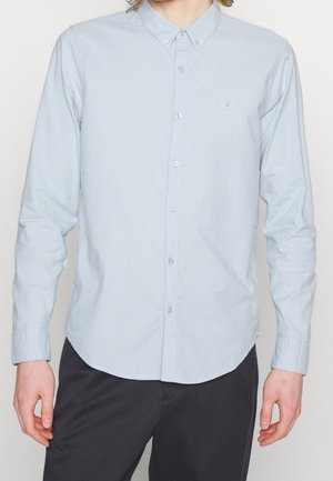 Chemise à manches longues bleu clair avec col pointu, ornée d'un petit logo sur la poitrine et d'un ourlet incurvé.