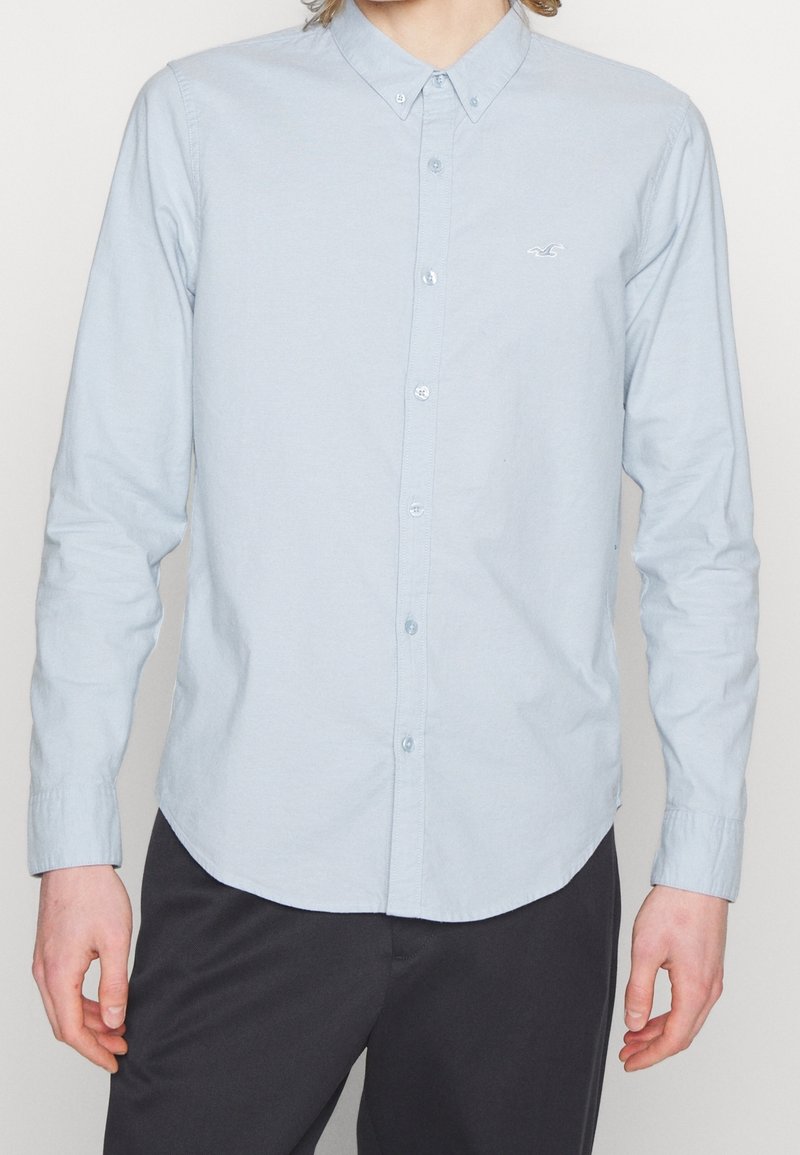 Ljust blå skjorta med lång ärm och knäppning, med spetsig krage, en liten logo på bröstet och en böjd sömdesign.