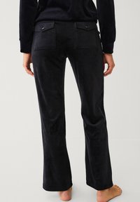 Pantalon noir évasé en matière douce et veloutée. Il présente deux poches arrière avec rabats et des détails de couture subtils.
