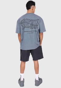 Graues oversized T-Shirt mit geschwungenem Textdesign auf dem Rücken, kombiniert mit schwarzen Shorts. Socken mit Text und graue Sneakers runden den Look ab.