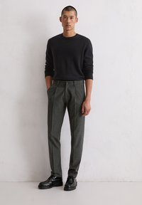 Grijze wollen broek met plooien en een knoopsluiting, gecombineerd met een zwarte trui en zwarte schoenen, tegen een neutrale muur als achtergrond.