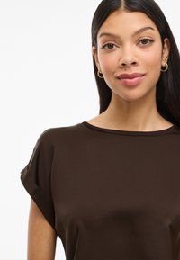 Brūns īsām piedurknēm tops ar gludu tekstūru un apaļu kakla izgriezumu. Mīksta auduma maigi pieglausta pie pleciem. Nav ievērojamu rakstu vai akcentu.