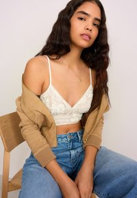 Witte kanten bralette met verstelbare bandjes, gecombineerd met een tan kleurige zip-up jas en high-waisted blauwe spijkerbroeken. Zichtbare geribbelde manchetten op de jas.