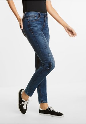 Jeans de mezclilla azul con un corte ajustado, que presentan bolsillos de parche y detalles desgastados. Combinados con zapatillas negras y una camiseta negra.