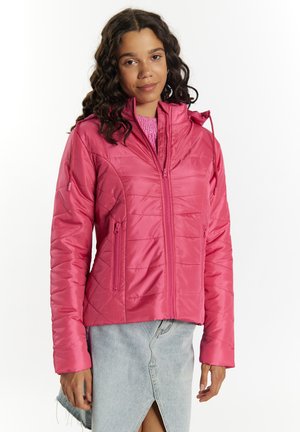 Ung kvinde med mørkt krøllet hår iført en knaldpink quiltet jakke og lyseblå denim nederdel med frontslids mod en hvid baggrund.