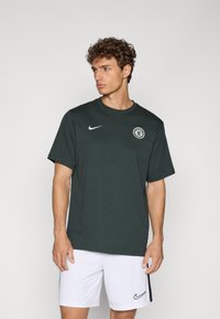 Donkergroen Nike T-shirt met korte mouwen, ronde hals en een cirkelvormig logo op de borst. Model draagt witte shorts met een zwart accent.