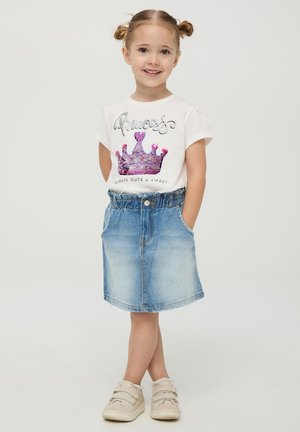 T-shirt bianco con grafica di una corona metallica, abbinato a una gonna di jeans con vita elastica e tasche. Sneakers di colore chiaro completano il look.
