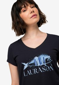 Černé tričko s výstřihem do V má modrou grafiku ryby a slovo "LAURASØN" ve světle modré barvě, volný střih a krátké rukávy.