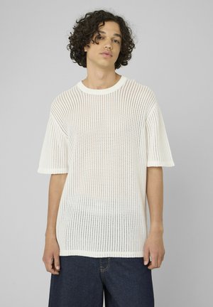 Jeune personne aux cheveux bouclés portant un pull en maille blanc à manches courtes perforé et un jean foncé sur un fond gris uni.