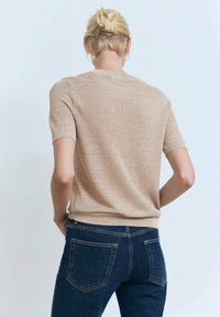 Beige strikket sweater med tekstur og korte ærmer, ribbet kant, båret med mørkeblå jeans, set fra ryggen mod en lys baggrund.