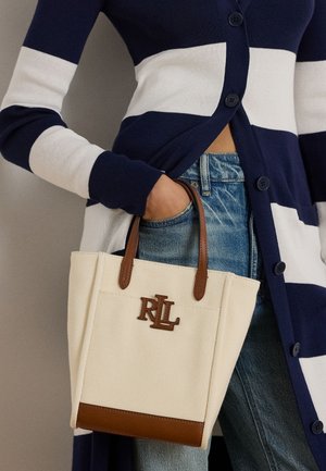 Persoană care poartă un cardigan cu dungi albastre și albe și blugi, ținând o geantă tote crem cu mânere maro și logo-ul RLL.