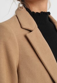 Kamelfarvet, tekstureret uldfrakke med en spids krave, båret over en sort ribbet turtleneck og parret med en sølvfarvet hoop ørering.