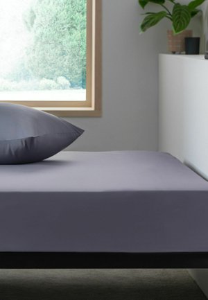Einzelbett mit grauem Spannbettlaken und Kissen neben einem Fenster, das grüne Bäume zeigt, ein weißer Schrank mit Pflanzen in einem minimalistischen Schlafzimmer.