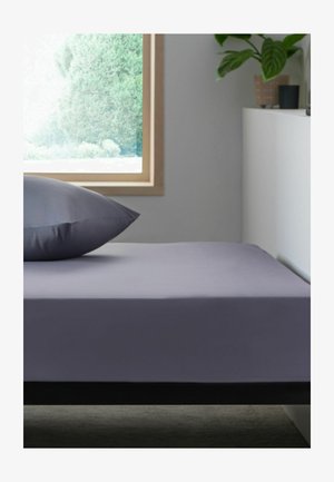 Einzelbett mit grauem Spannbettlaken und Kissen neben einem Fenster, das grüne Bäume zeigt, ein weißer Schrank mit Pflanzen in einem minimalistischen Schlafzimmer.