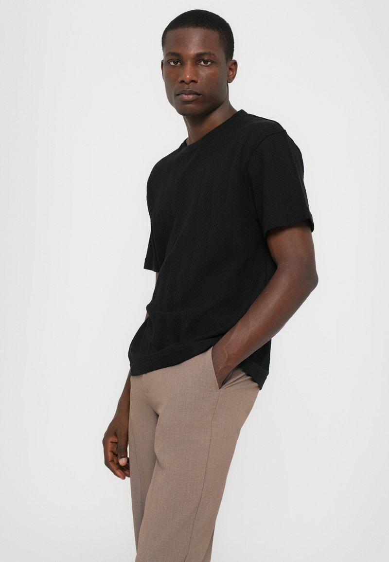 Black short-sleeve t-shirt with a textured herringbone pattern, paired with light brown tailored trousers, worn with relaxed posture.