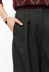 Pantalon gris sur mesure avec une poche avant, présentant des plis et une texture lisse. Une main est visible, ornée d'un bracelet circulaire.