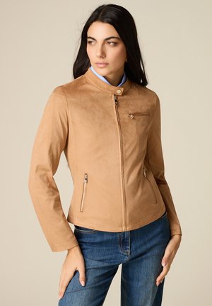 Frau mit langen dunklen Haaren, die eine hellbraune Wildlederjacke mit Reißverschlusstaschen und blaue Jeans trägt, schaut seitlich vor einem beigen Hintergrund.