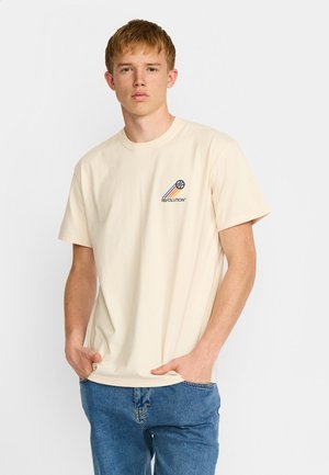 Jeune homme portant un T-shirt crème avec un petit motif de basket-ball et le texte "REVOLUTION", associé à un jean bleu, debout les mains dans les poches.