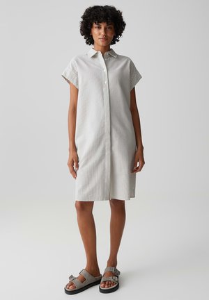 Frau mit lockigem Haar, die ein knielanges, kurzärmliges, gestreiftes Kleid mit Knöpfen und graue Sandalen mit Schnallen trägt, steht vor einem schlichten Hintergrund.