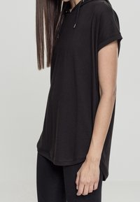 Svart kortärmad hoodie med en lös passform, dragsko i halsen och rundad nederkant, presenterad mot en neutral bakgrund.