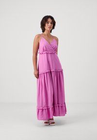 Rochie lungă roz cu un design înfășurat, accente volan, și bretele subțiri. Textură moale și model evazat în partea de jos, asociată cu încălțăminte roz deschis.