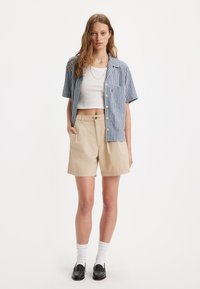Chemise à manches courtes en vichy bleu et blanc, shorts beiges, chaussettes blanches, mocassins noirs, design minimaliste avec une poche sur la chemise.