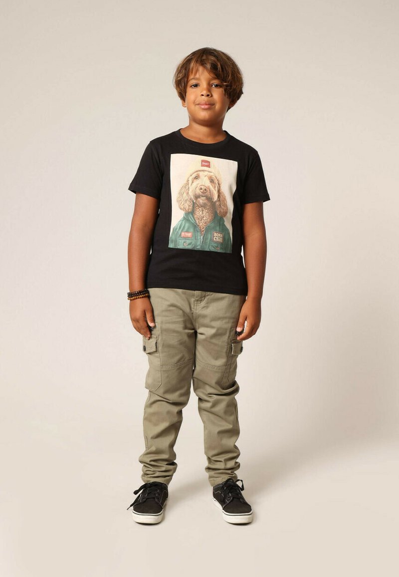 T-shirt noir avec un graphisme d'un chien en tenue verte. Un pantalon cargo olive avec des poches et des baskets noires complètent la tenue.