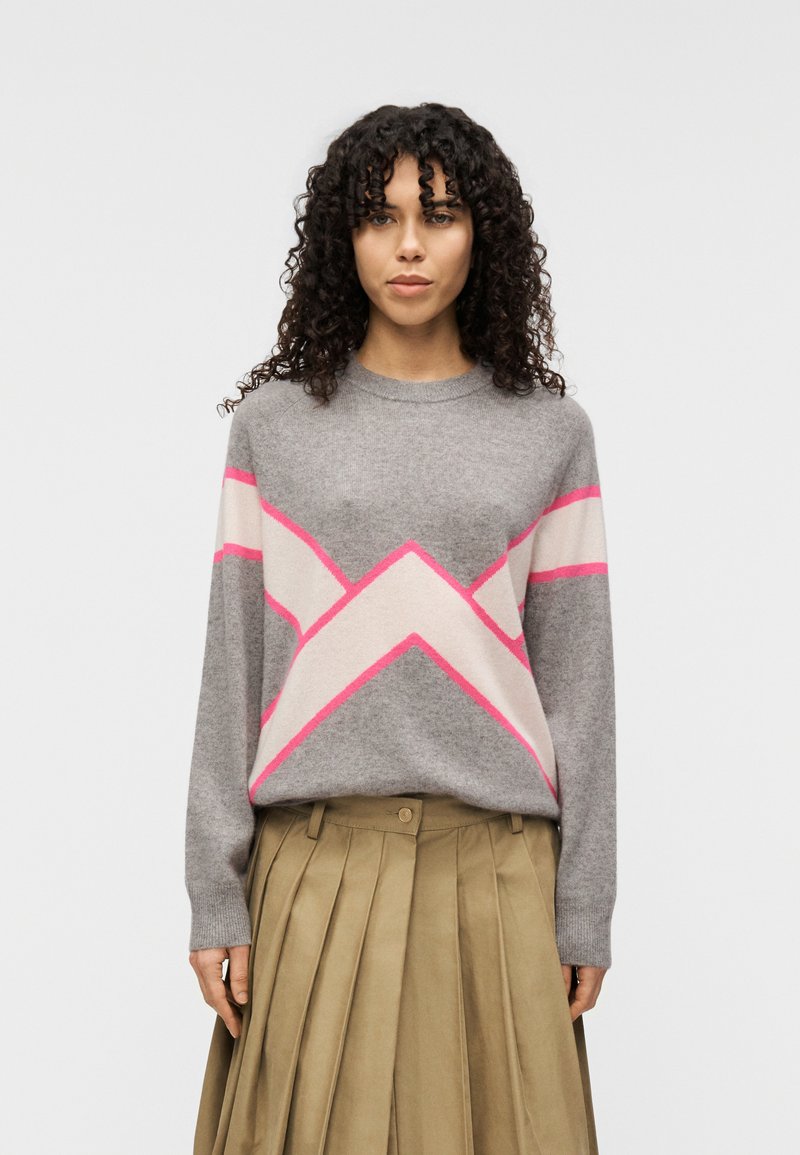 Pull gris avec motifs en zigzag rose et crème, col rond, manches longues ; associé à une jupe plissée kaki. Matière tricotée douce et texturée.