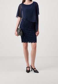 Robe de soirée marine avec superposition en tissu transparent, ornée de paillettes et à ourlet festonné. Associée à une petite pochette métallique et des escarpins noirs à bride cheville.