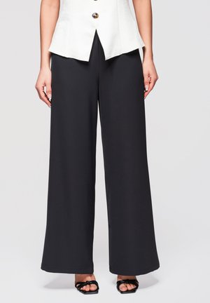 Femme portant un pantalon large noir, un top blanc sans manches boutonné, et des sandales noires à brides, se tenant devant un fond uni.