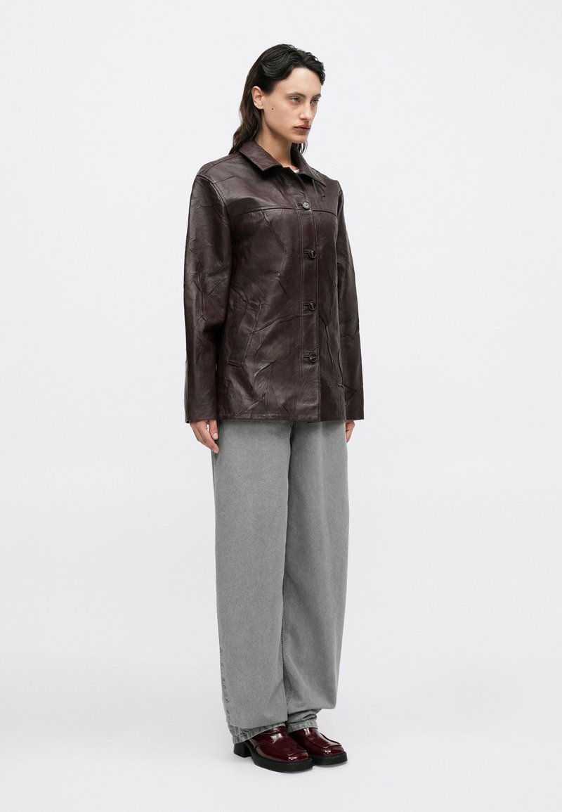 Veste en cuir marron avec col, fermeture à boutons sur le devant et finition texturée, associée à un pantalon gris à jambes larges et des chaussures rouge foncé.