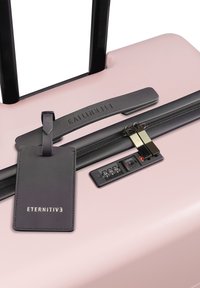 Valigia rigida di colore rosa chiaro con manico telescopico nero, cerniera con doppi cursori in metallo e un'etichetta per bagagli nera con testo in rilievo.