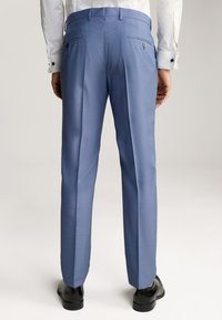 Pantalons de costume bleu avec une coupe plate, ajustement sur mesure, deux poches arrière à boutons et un design élégant, associés à des chaussures noires.