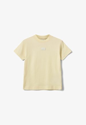 T-shirt à manches courtes de couleur crème avec un col rond et un petit patch rectangulaire centré sur la poitrine.