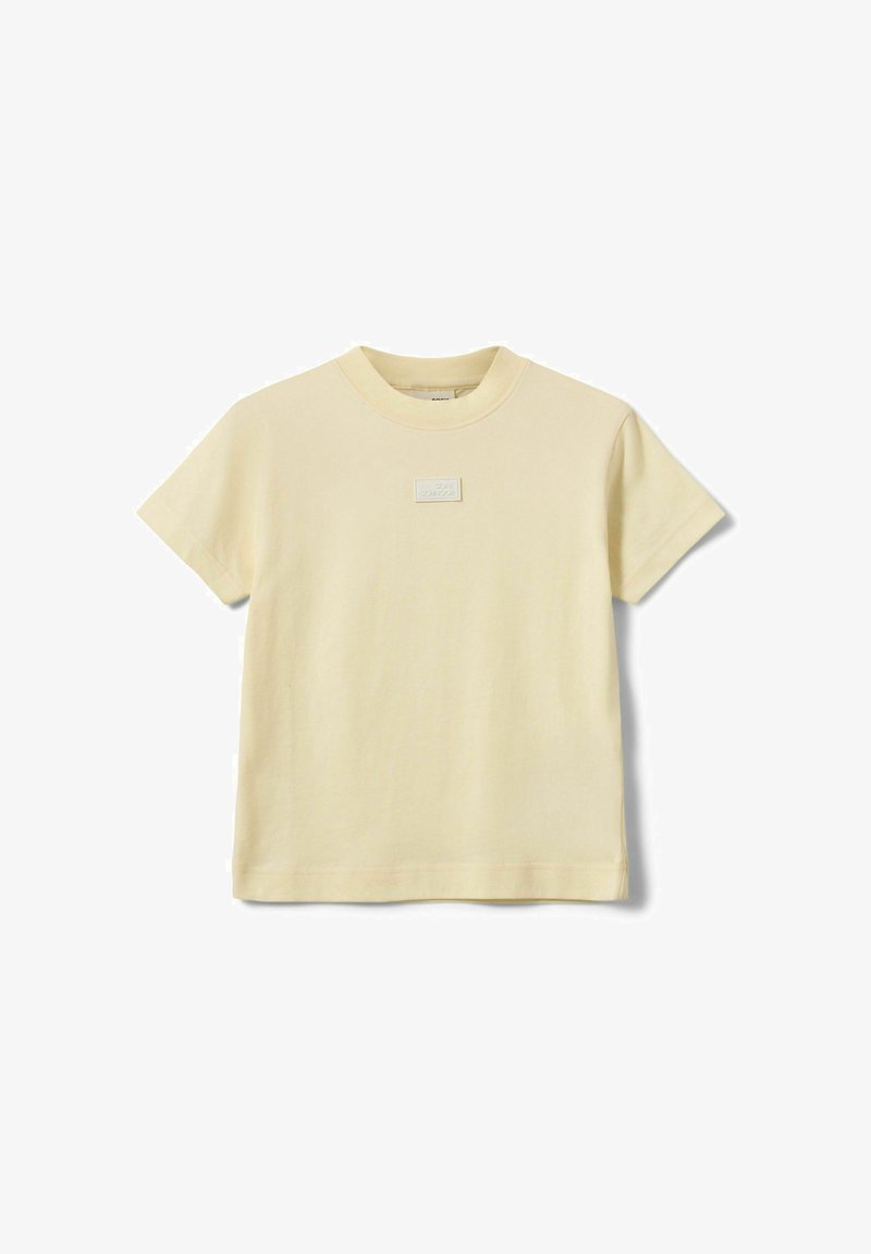 T-shirt à manches courtes de couleur crème avec un col rond et un petit patch rectangulaire centré sur la poitrine.