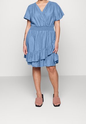 Femme portant une robe portefeuille bleue longueur genoux avec un ourlet volanté et une taille smockée, associée à des sandales roses tissées à talons.