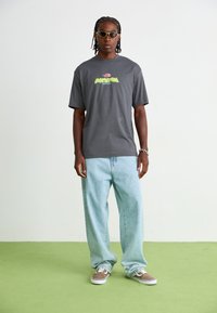 Grå kortærmet t-shirt med farverigt grafisk motiv, parret med lyseblå baggy jeans og brune sneakers. Accessories inkluderer lyse solbriller.