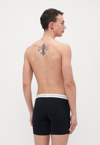 Čierne boxerky s bielym pásom "COPENHAGEN STUDIOS". Hladká tkanina, priliehavý strih. Model zobrazený zozadu, s viditeľným tetovaním.
