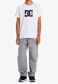Bílá bavlněná tričko s černým logem DC, spárované se šedými volnými džínami a černými teniskami s bílými akcenty.