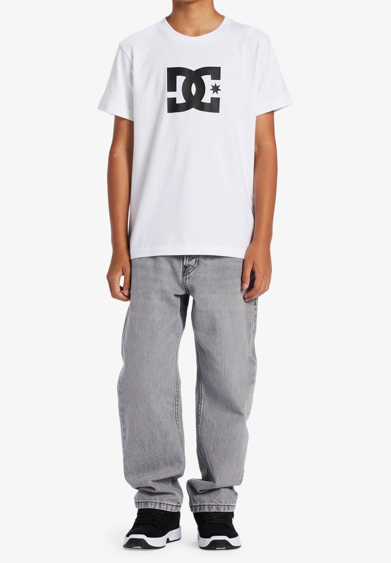 Bílá bavlněná tričko s černým logem DC, spárované se šedými volnými džínami a černými teniskami s bílými akcenty.