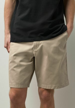 Férfi, bézs, térdig érő rövidnadrágban és fekete pólóban, egyik kezével a zsebében, semleges háttér előtt.